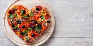 Baked heart-shaped homemade pizza on a cutting board on white wooden background. Close-up